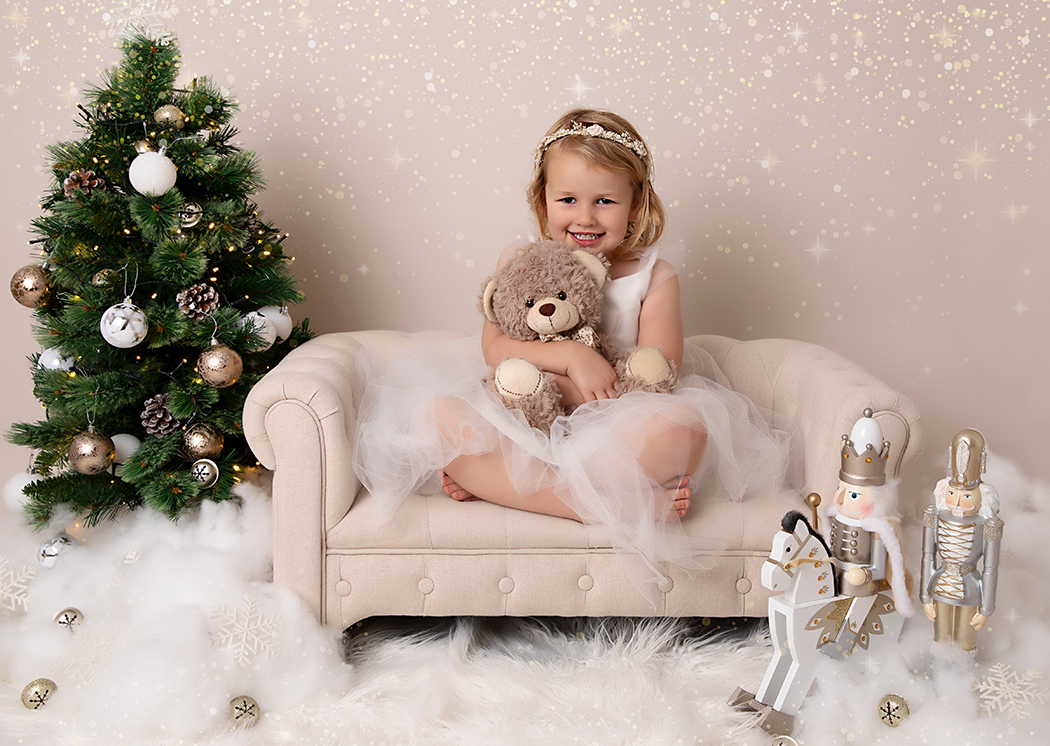 3 year old girl at her Christmas photoshoot sat on a miniature sofa cuddling her favourite teddy bear